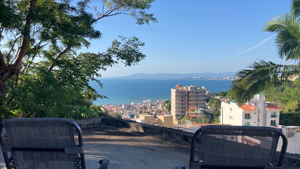Deux chaises sont placées face à un paysage pittoresque d’une ville côtière, avec ses bâtiments et un ciel dégagé.