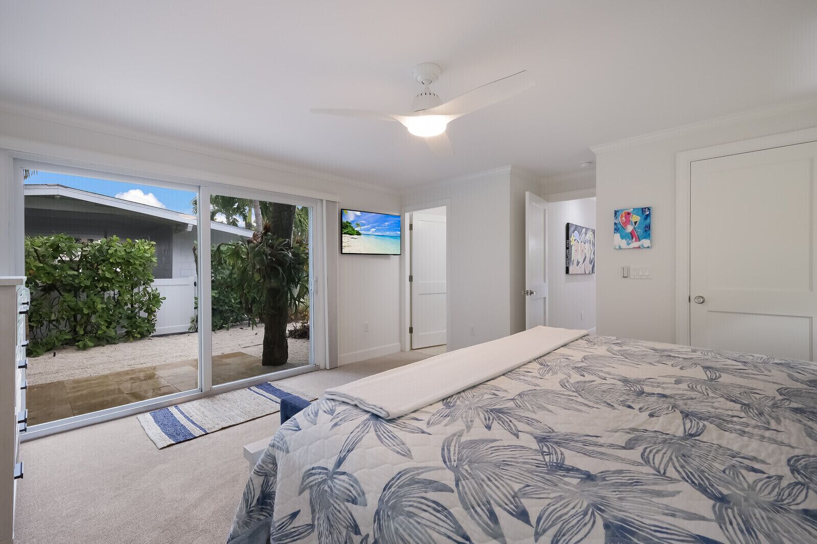 A bedroom with a bed, a ceiling fan, a TV, and a sliding glass door leading to a balcony with a view of the beach.