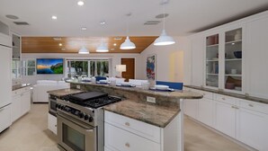 A modern kitchen with white cabinetry, a granite countertop, stainless steel appliances, pendant lighting, and a view of a tropical landscape through the windows. - Canal Walk: Tiki Bar, Mini Golf, Pool & Spa On Canal by Anna Maria Life Vacation Rentals (Anna Maria)