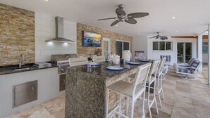 A modern kitchen with granite countertops, a built-in stove, and a stone backsplash. There are bar stools and a dining table with a TV above it. - Canal Walk: Tiki Bar, Mini Golf, Pool & Spa On Canal by Anna Maria Life Vacation Rentals (Anna Maria)