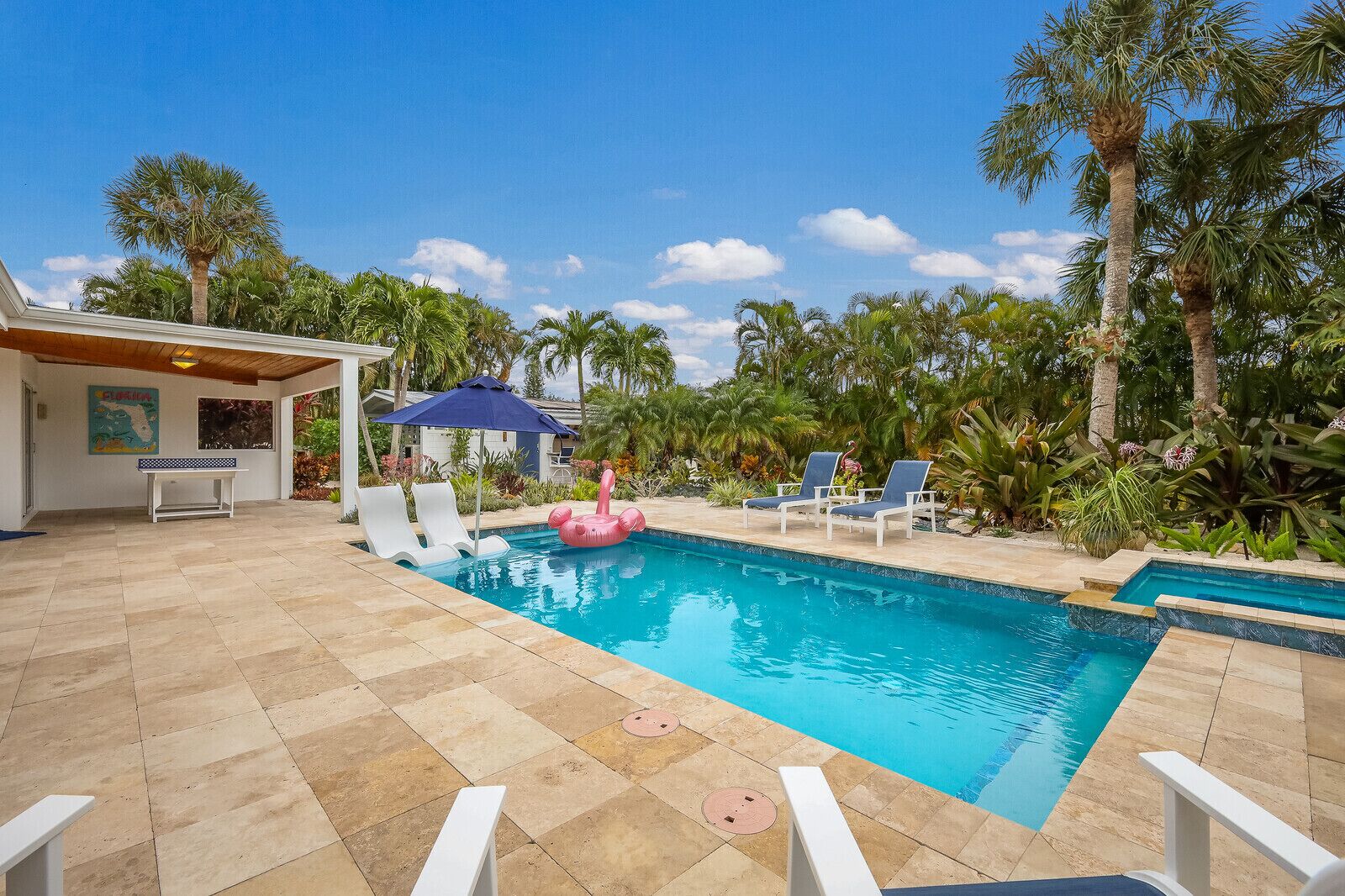 A swimming pool with lounge chairs and a pink inflatable float, surrounded by palm trees and a covered patio area.