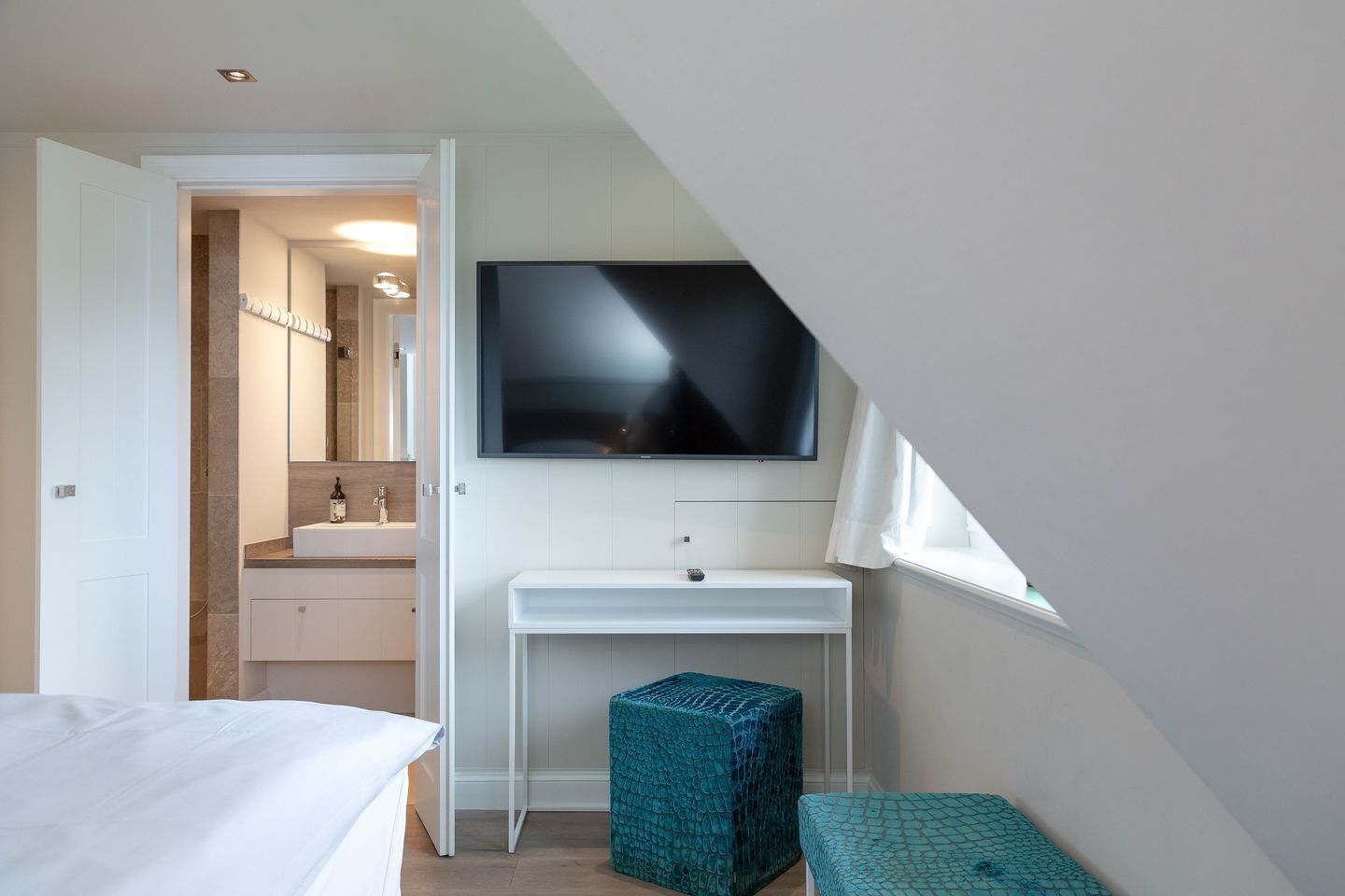 A bedroom with a bed, a TV mounted on a wall, a small white desk, and a window.