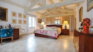 Une chambre avec un plafond en bois, un lit recouvert d’une couvre-lit à motifs floraux, deux tables de chevet avec des lampes, une commode et une chaise.