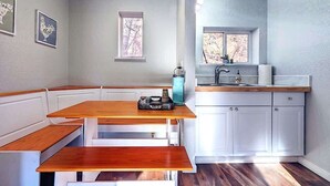 Una cocina con mesa y banco de madera, armarios blancos, un ventanal y un lavamanos.