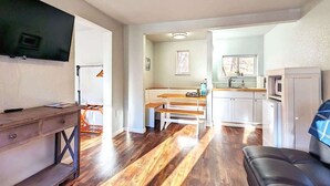 Una sala de estar con televisor, un escritorio de madera, un banco, un sofá y una zona de cocina con ventana.