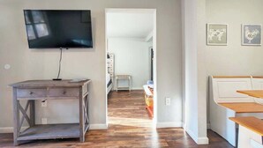 Una sala de estar con televisor, un escritorio de madera, un banco y una puerta que da a otra habitación.