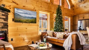 Een gezellig houten blokhutinterieur met een houten wand, een grote televisie, een kerstboom, een leren bank en een eettafel met eten en drinken.