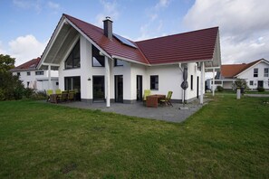 Una casa moderna de dos pisos con techo inclinado, ventanas grandes y un patio con mobiliario exterior.