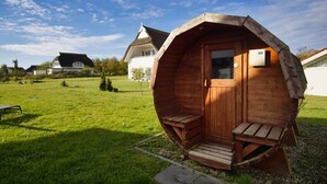 Eine hölzerne Geodätkuppel mit einer Tür und zwei Bänken, eingebettet in eine grüne Wiese, mit Häusern im Hintergrund.