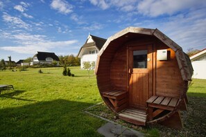 Une structure en forme de dôme géodésique, faite de bois, avec une porte et deux bancs, située dans une zone herbeuse, avec des maisons en arrière-plan.
