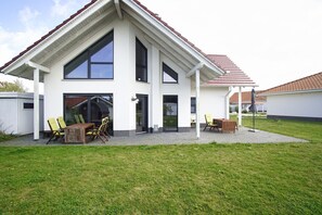 Moderni, kaksikerroksinen talo, jossa on katettu terassi, suuret ikkunat ja grillauspaikka.