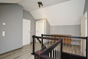 Ein geräumiges Zimmer mit Holzboden, einer dunklen Holztreppe mit Metallgeländer und einer Deckenleuchte.