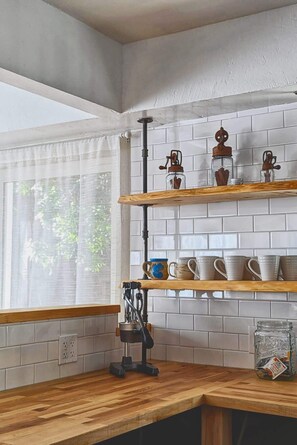 Una cocina con paredes de azulejos blancos, estantes de madera y una ventana con cortinas de gasa.