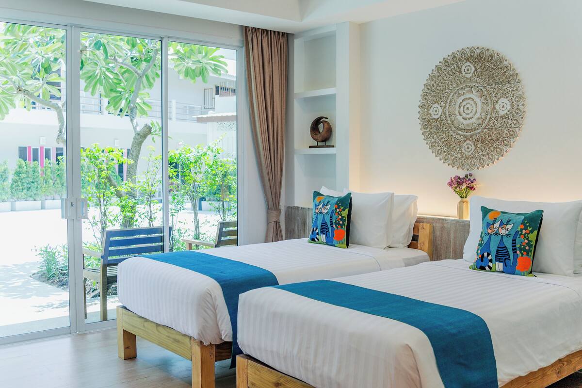 Une chambre d’hôtel avec deux lits, une grande fenêtre donnant sur un paysage verdoyant, un élément décoratif mural et un rideau.
