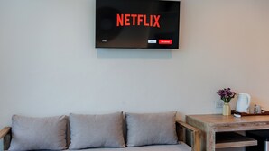 Ένα σαλόνι με μια τηλεόραση που προβάλλει το λογότυπο της Netflix, έναν καναπέ και ένα ξύλινο βοηθητικό τραπέζι με ένα βάζο και μια καφετιέρα.