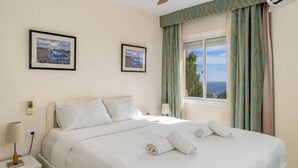 Una habitación de hotel con una cama, dos cuadros en la pared, una mesita de noche con una lámpara y una ventana con cortinas.