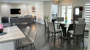 Una sala de estar moderna con un televisor de pantalla plana, un comedor con mesa de vidrio y una cocina con encimera blanca.