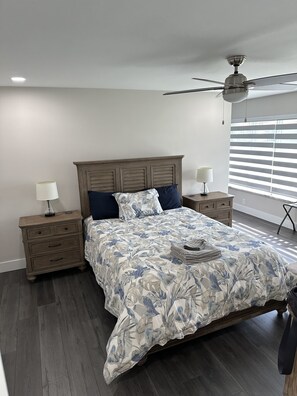 Une chambre à coucher comprenant un lit, deux tables de chevet, un ventilateur de plafond et une fenêtre avec des stores.