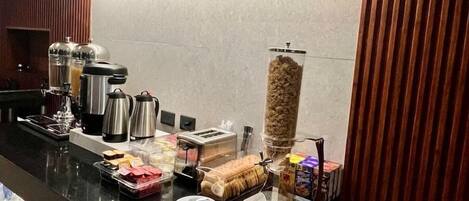 Una barra de desayuno con máquinas de café, una jarra de café, un dispensador de cereales y diversos productos para el desayuno.