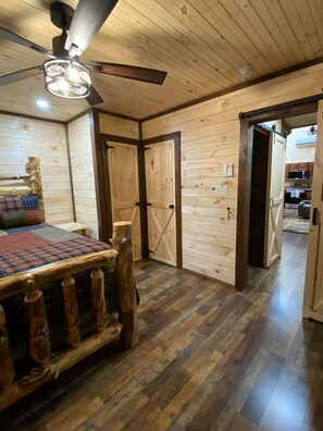 Ein Holzblockhaus-Innenraum mit einem Bett, einem Deckenventilator und Blick in die Küche.
