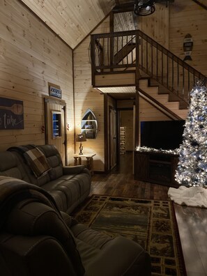 A cozy cabin interior with a leather sofa, a Christmas tree, and a staircase leading to an upper level.
