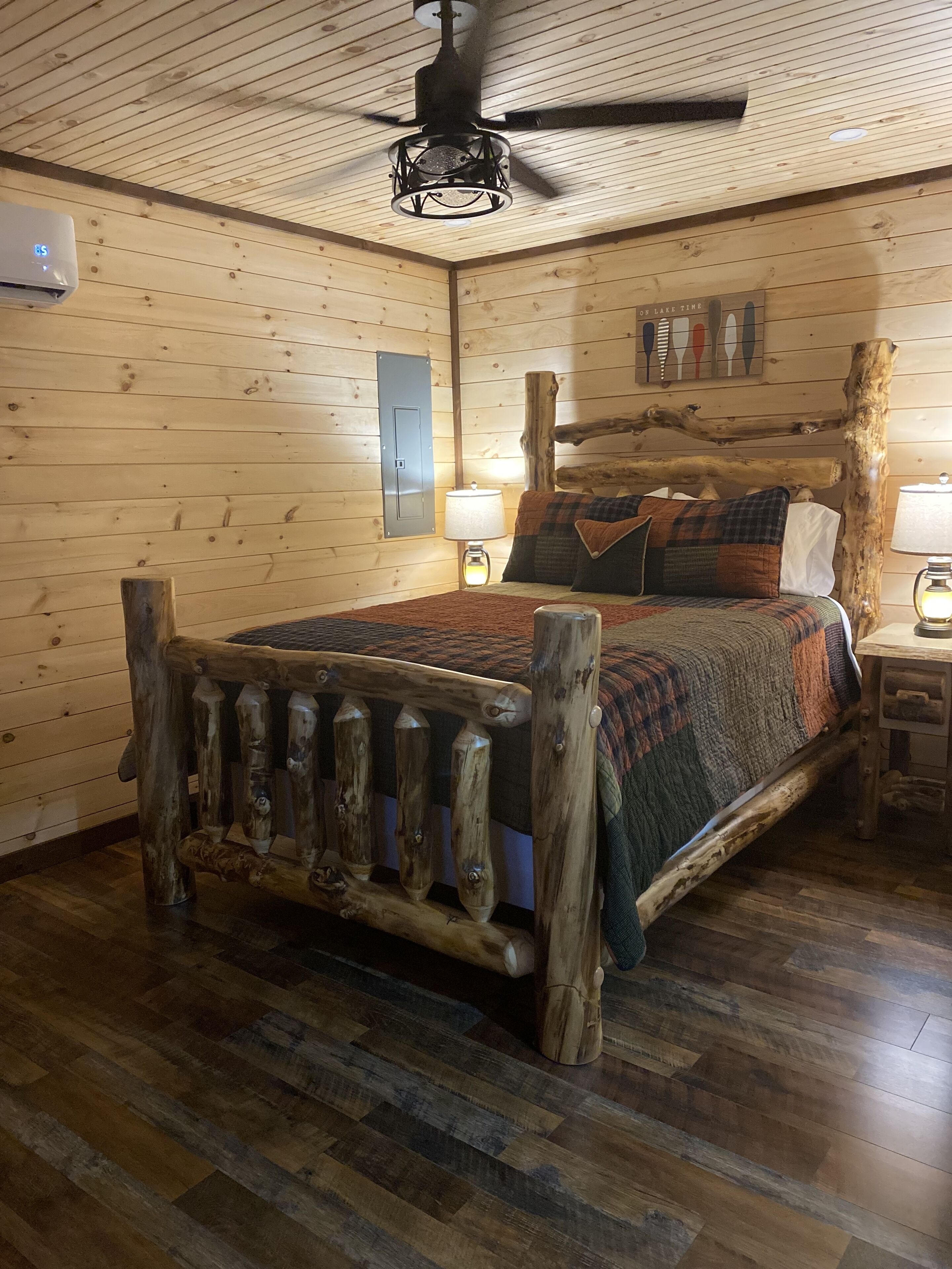 A rustic wooden room with a bed, wooden walls, a ceiling fan, and a nightstand with a lamp.