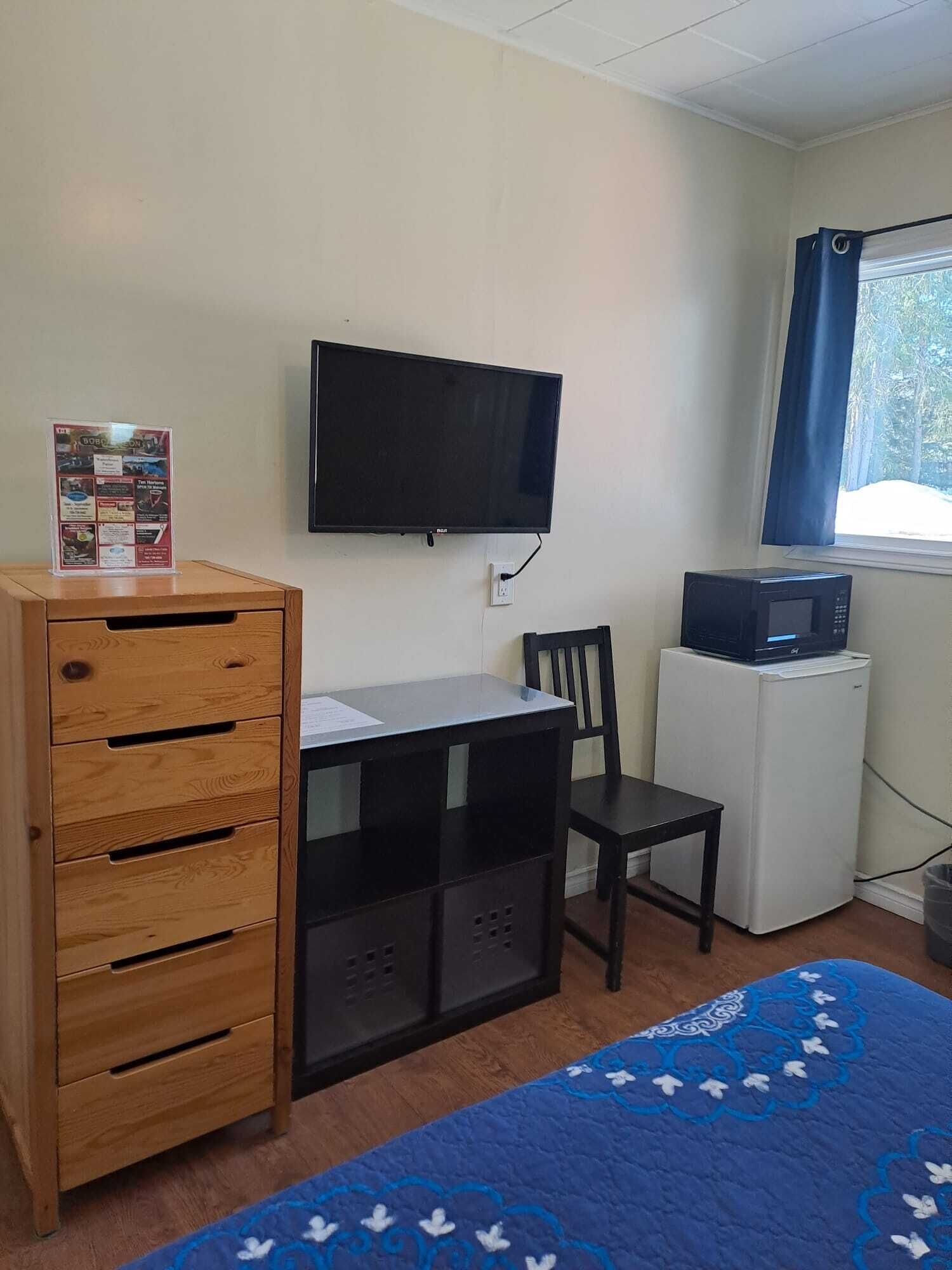 Une petite pièce comprenant un meuble en bois, un bureau, une chaise, un téléviseur, un four à micro-ondes et une fenêtre avec des rideaux.