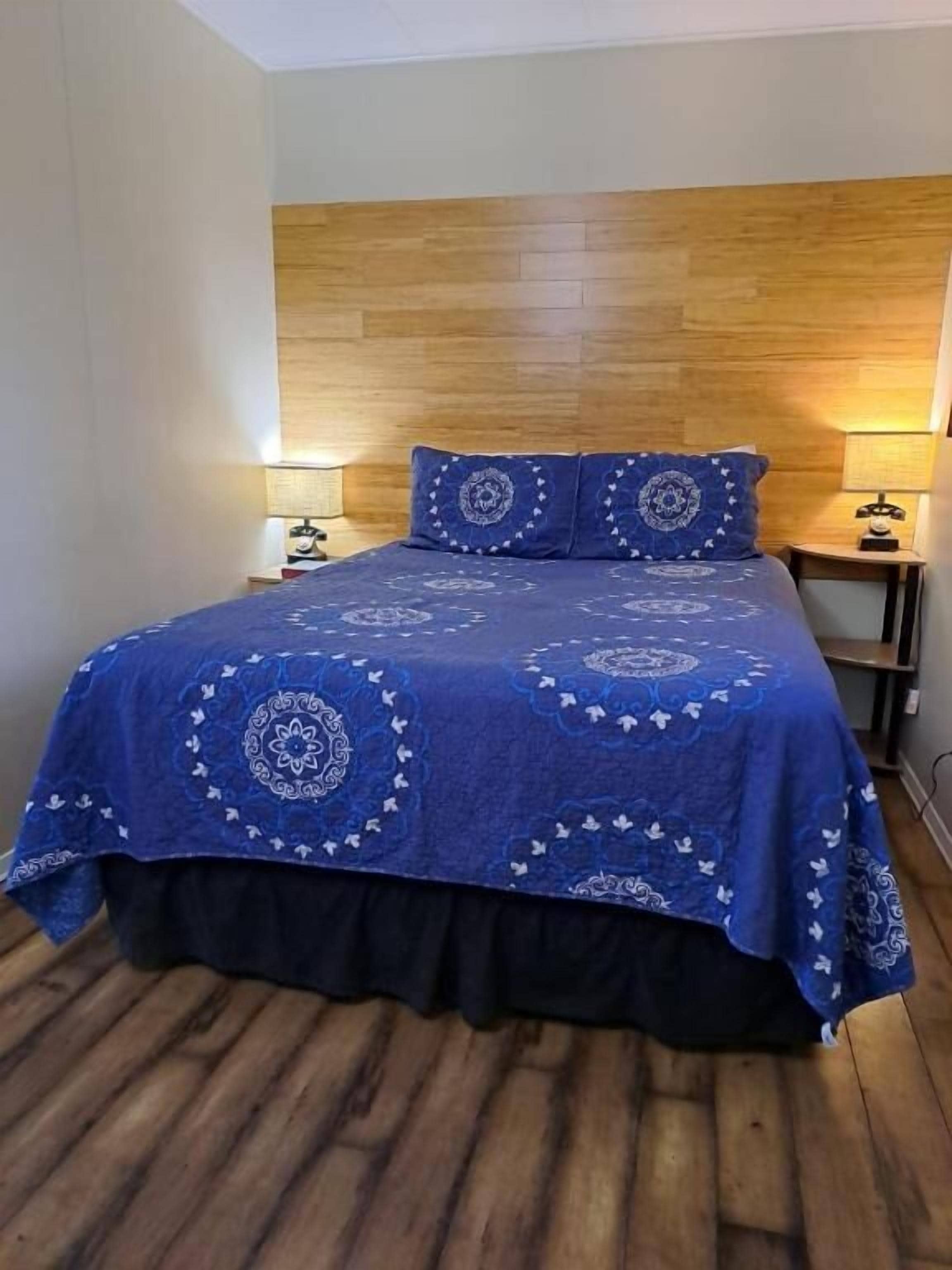 Una habitación con cabecera de madera, cama con colcha azul adornada con motivos circulares, dos lámparas de noche y suelo de madera.