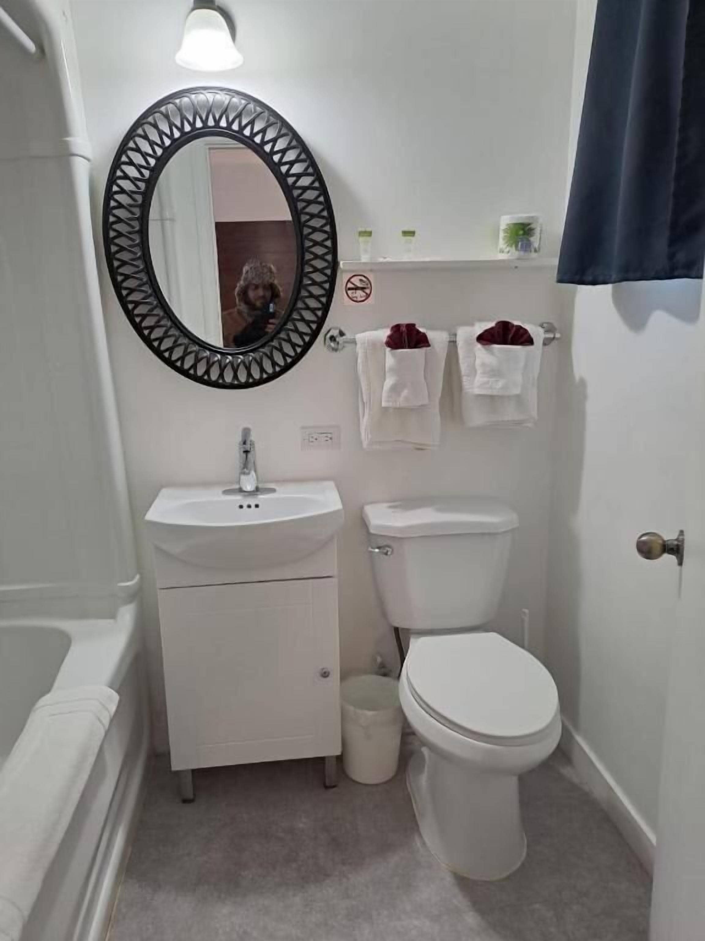Un baño pequeño y funcional con lavamanos, inodoro y bañera blancos. Se observa un espejo redondo con un marco decorativo, además de un perchero con dos toallas.