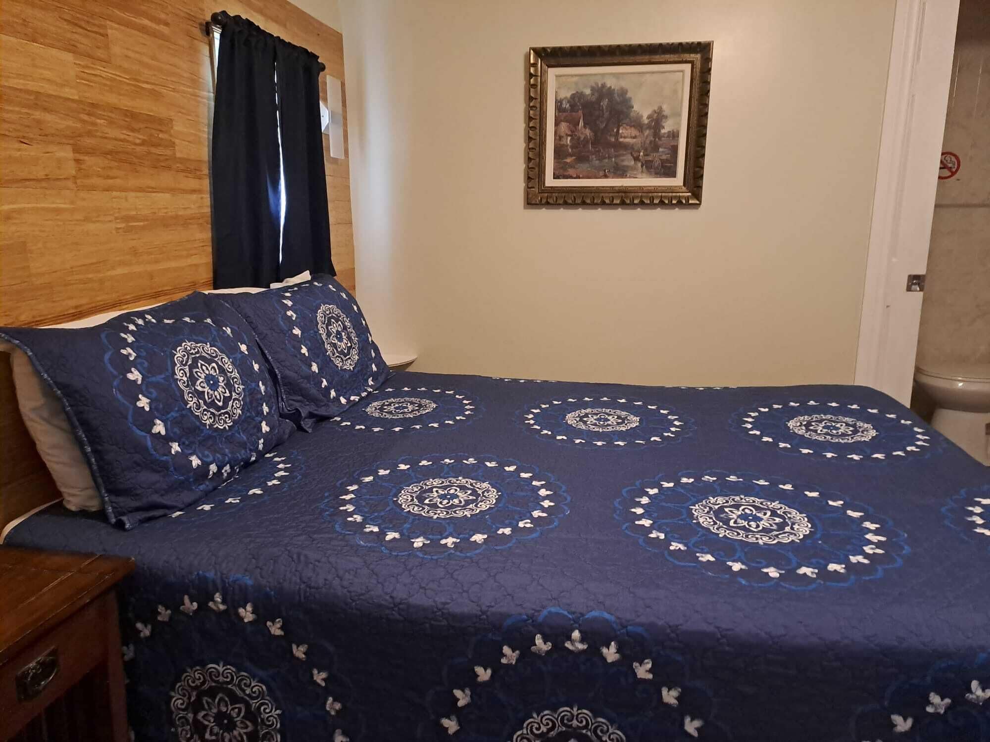 Quarto com cama coberta por um edredom azul com desenhos brancos, cabeceira de madeira, quadro emoldurado na parede e porta com placa de proibido fumar.