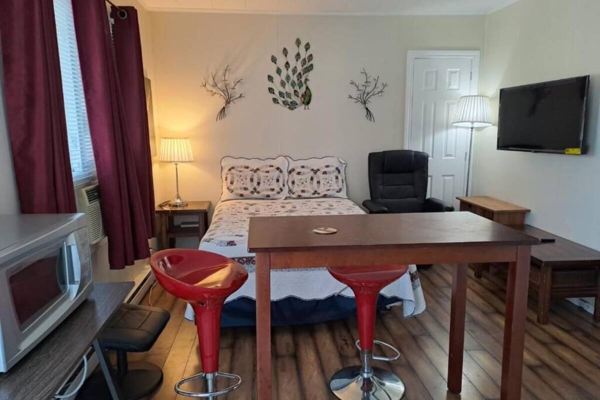 A hotel room with a bed, two red bar stools, a wooden table, a TV, a lamp, and a decorative wall piece.