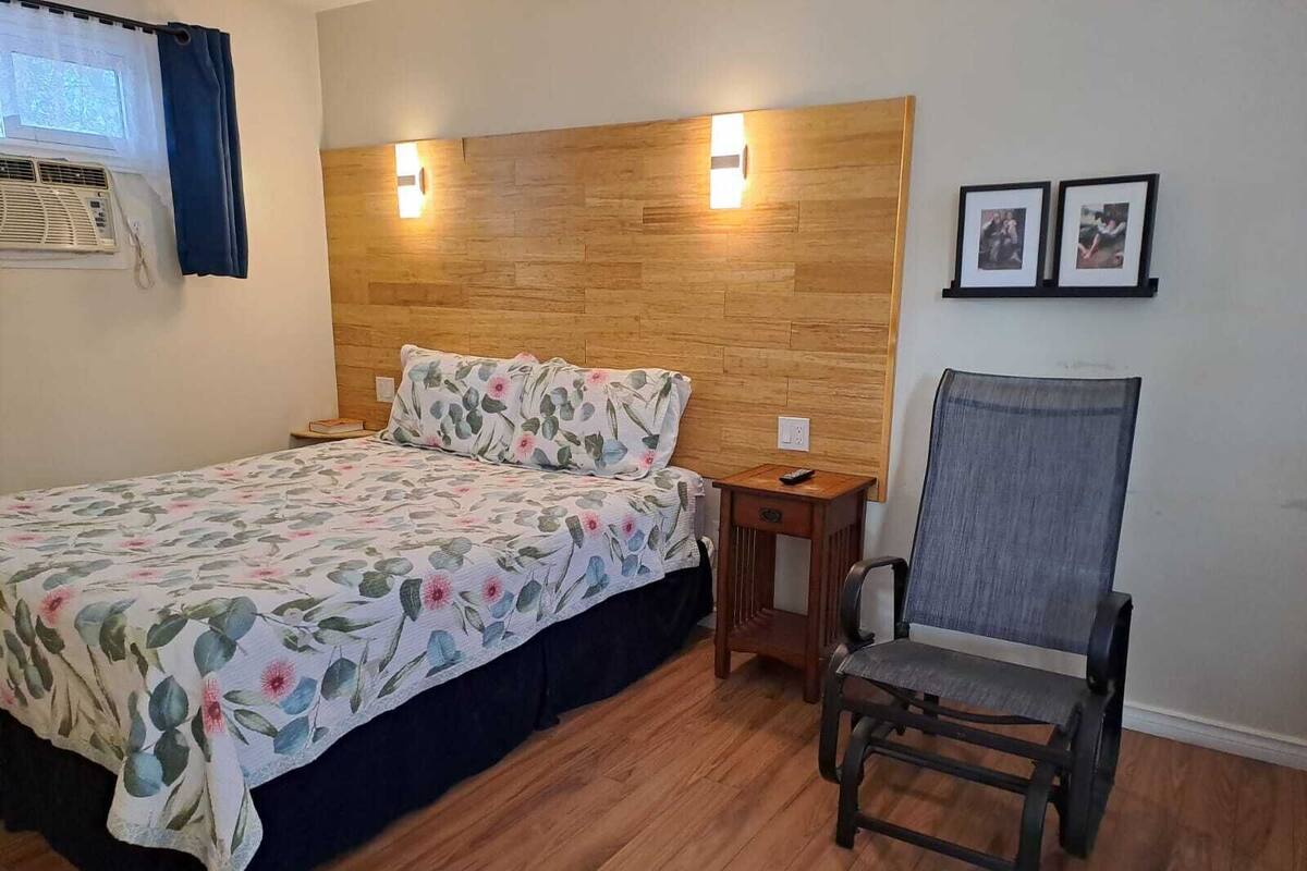 A bedroom with a wooden headboard, a floral bedspread, a wooden nightstand, a chair, and two framed pictures on the wall.