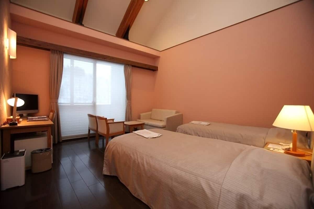 Ein Hotelzimmer mit zwei Betten, einem Schreibtisch mit Computer, einem Stuhl, einem kleinen Tisch und einem Fenster mit Vorhängen.