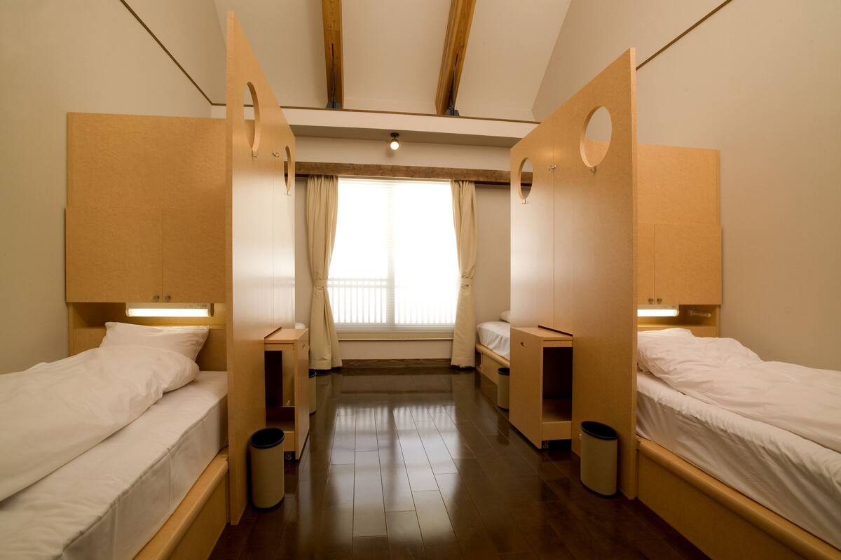 Ein Zimmer mit zwei Einzelbetten, Holz-Kopfteilen, einem kleinen Tisch und einem Fenster mit Vorhängen.