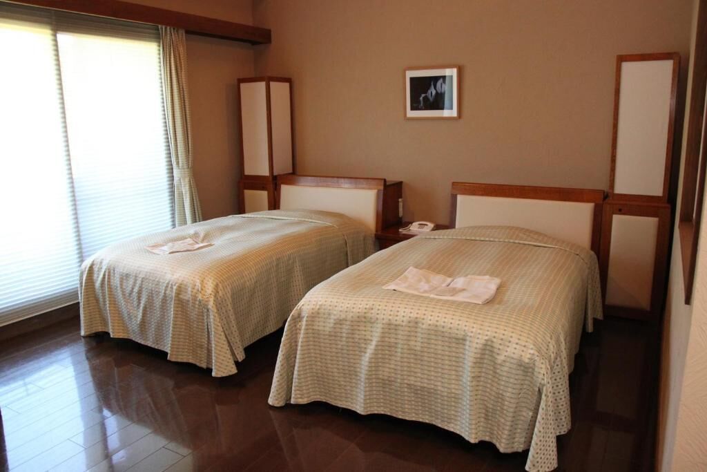 A hotel room with two single beds, a bedside table with a lamp, a framed picture on the wall, and a window with curtains.