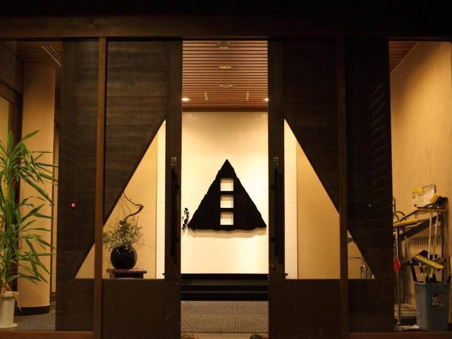 A traditional Japanese-style entrance with a wooden structure, a black triangular decoration on the wall, and a potted plant to the left.