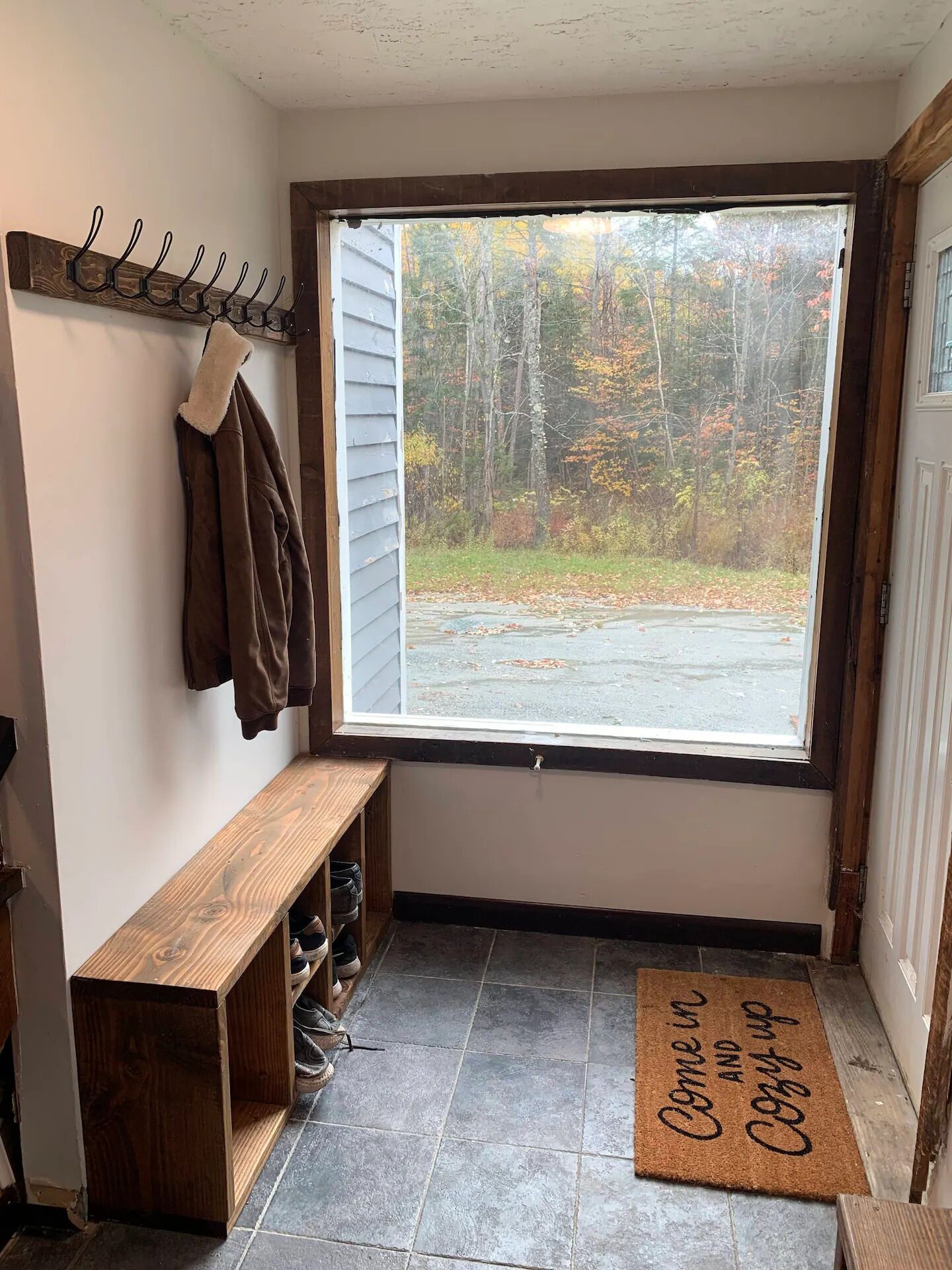 Un petit hall d’entrée avec un banc, un porte-manteau et une vue sur une zone boisée par une fenêtre.