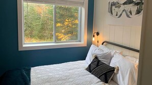 Een slaapkamer met een groot raam, een bed met wit beddengoed, een zwart-wit kussen en een wandlamp.