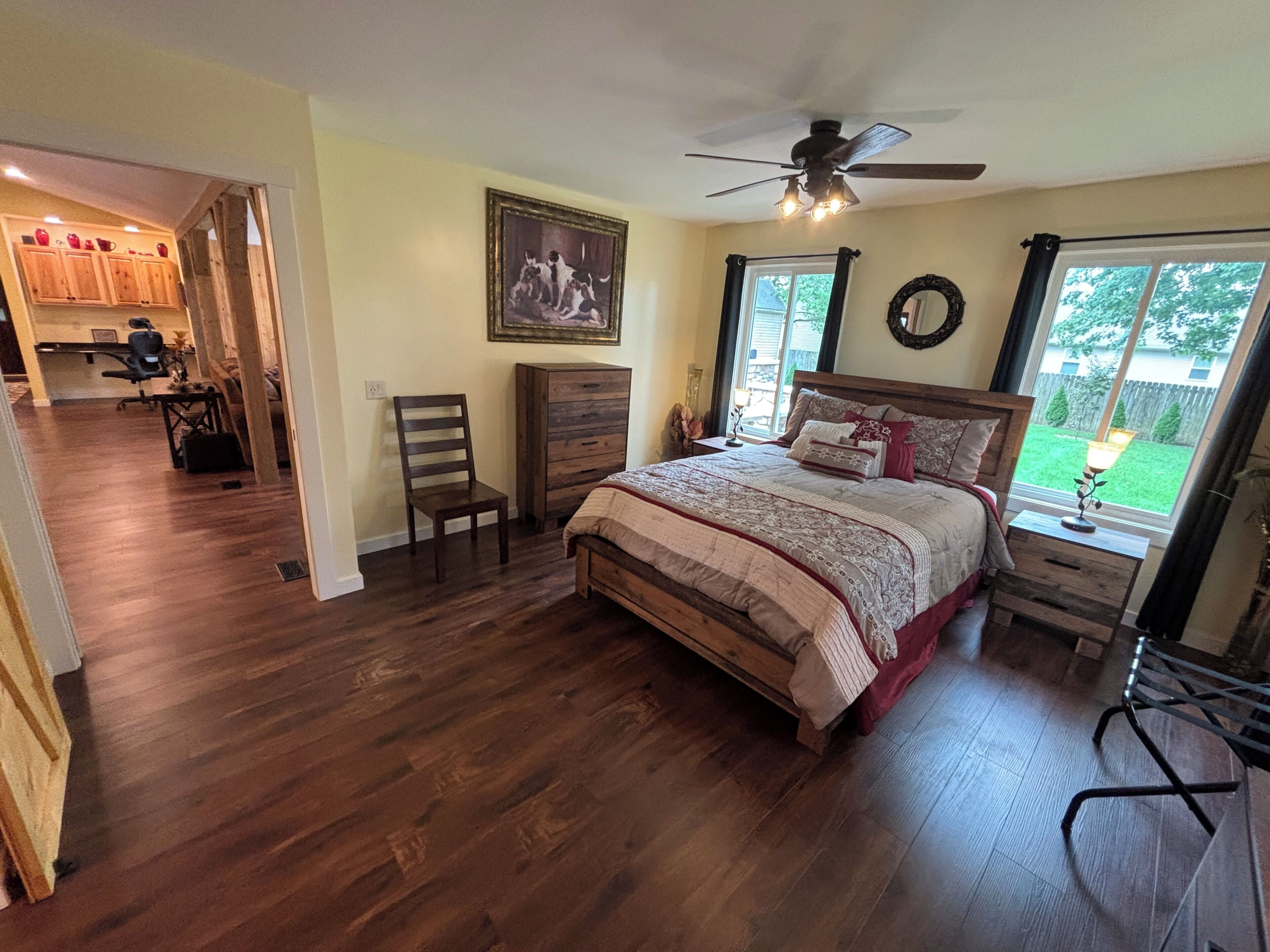 A bedroom with a bed, a ceiling fan, a window with a view of the outdoors, and a wooden floor.