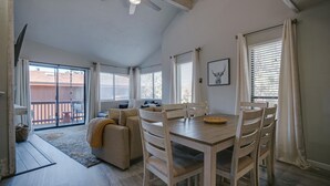 A spacious living area with a wooden dining table, chairs, and a sofa. There is a balcony with a sliding door and a ceiling fan. - Condo within walking distance of the Chile Express (Angel Fire)