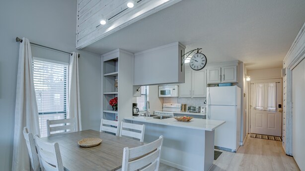 A modern kitchen with white cabinetry, a central island, and a dining area with a wooden table and chairs. - Condo within walking distance of the Chile Express (Angel Fire)