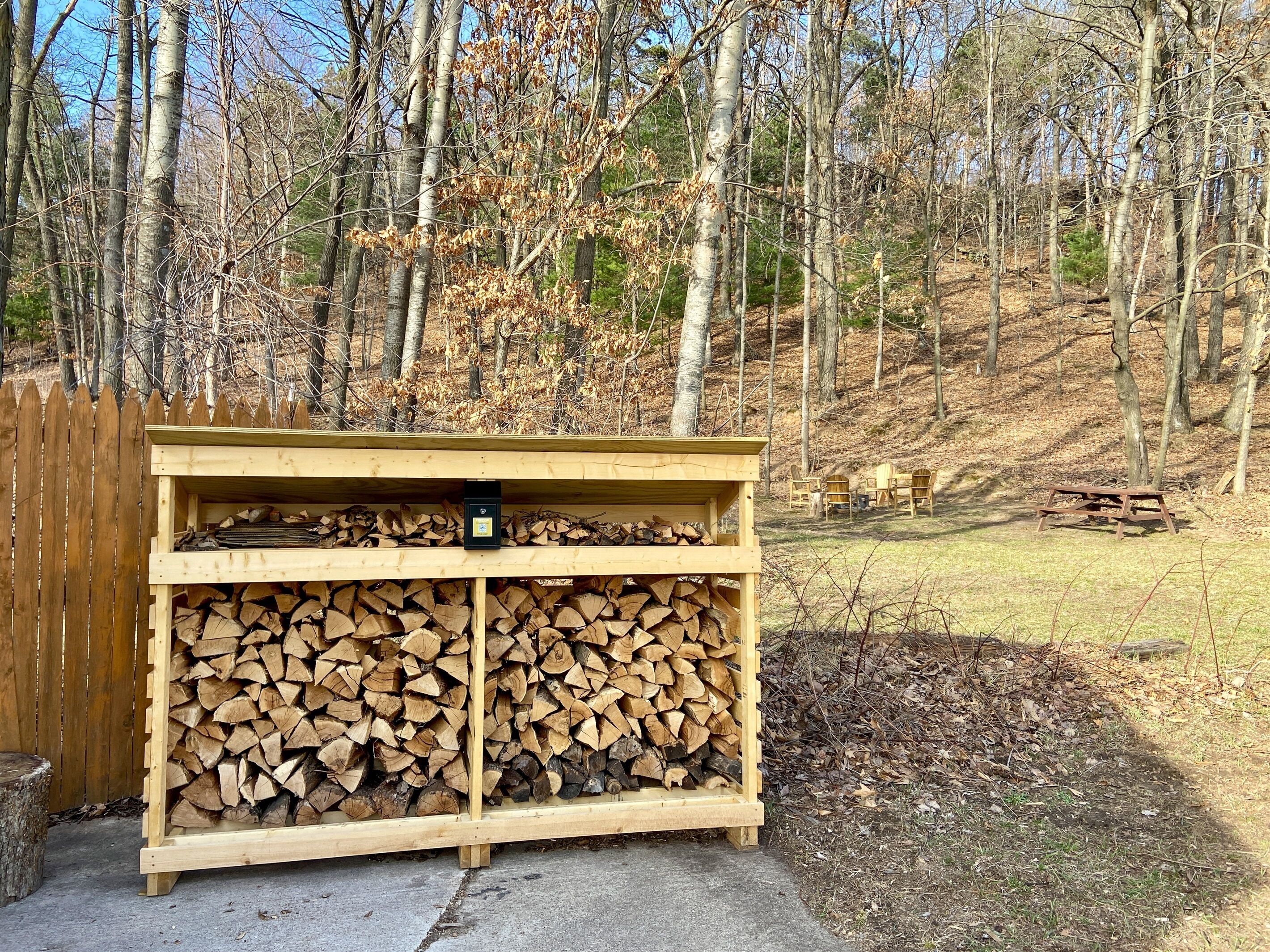 A wooden structure with two shelves of stacked firewood, a small black object on the top shelf, and a wooden fence in the background.