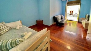 Ein Schlafzimmer mit einem Bett, einem kleinen Holzschemel, einem grauen Sofa und einem Balkon mit Geländer.