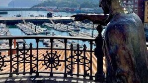 Eine Statue mit Blick auf einen Hafen, im Hintergrund Schiffe und Gebäude.