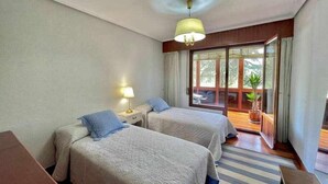 A hotel room with two beds, a nightstand with a lamp, a wooden desk, a striped rug, and a balcony with potted plants. - Amatxo Maitia - Large movie apartment. (Bilbao)