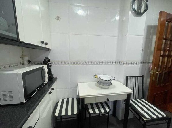 A small kitchen with white tiles, a microwave, a small table with a bowl, and two stools. - Goiz Etxea - Acogedor Piso en la Parte Antigua (Bermeo)