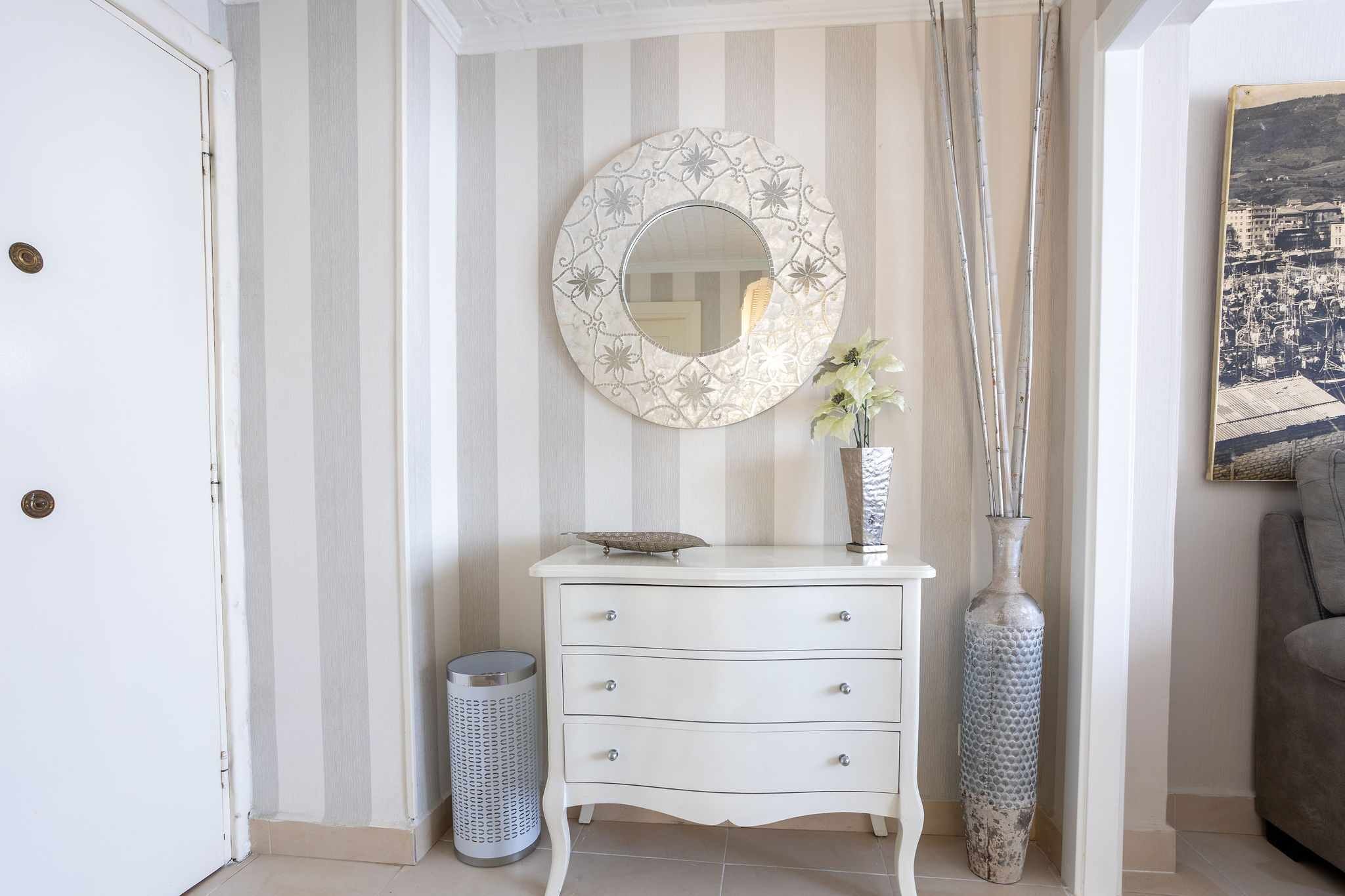 Een kamer met een witte dressoir, een ronde spiegel met bloemendecoratie, een vaas met bloemen en een ingelijst fotokader aan de muur.