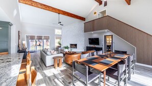 A spacious living area with a high ceiling, exposed wooden beams, a stone fireplace, a large TV, and a dining area with a wooden table and chairs.