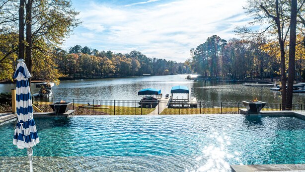 A swimming pool with clear water, surrounded by a deck with lounge chairs and umbrellas, overlooking a lake with boats and trees.