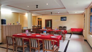 A dining area with multiple tables set for meals, wooden chairs, and a red tablecloth. - Ishwar Guest House (Dharamshala)